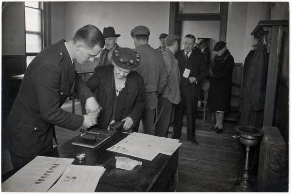 Archival black-and-white photograph, 1934, interior of Oskaloosa City Hall during a municipal fingerprint enrollment; silver gelatin print from a 4x5 Speed Graphic on tripod, panchromatic film with slight motion blur on hands, soft window light from high sash windows, uneven exposure falloff at frame edges, light paper curl and minor silvering at corners. Asymmetric composition with a dominant subject: a uniformed technician gently guiding an elderly woman's fingers across an ink roller at a waist-high table; behind them a loose queue of townspeople—farmers in work coats, a clerk in Sunday shoes—waits along a scuffed wooden floor. Background details include a coat rack, a spittoon, and a radiator; no legible signage or lettering anywhere. 127mm lens perspective, moderate depth of field, natural skin texture and varied ages/builds.