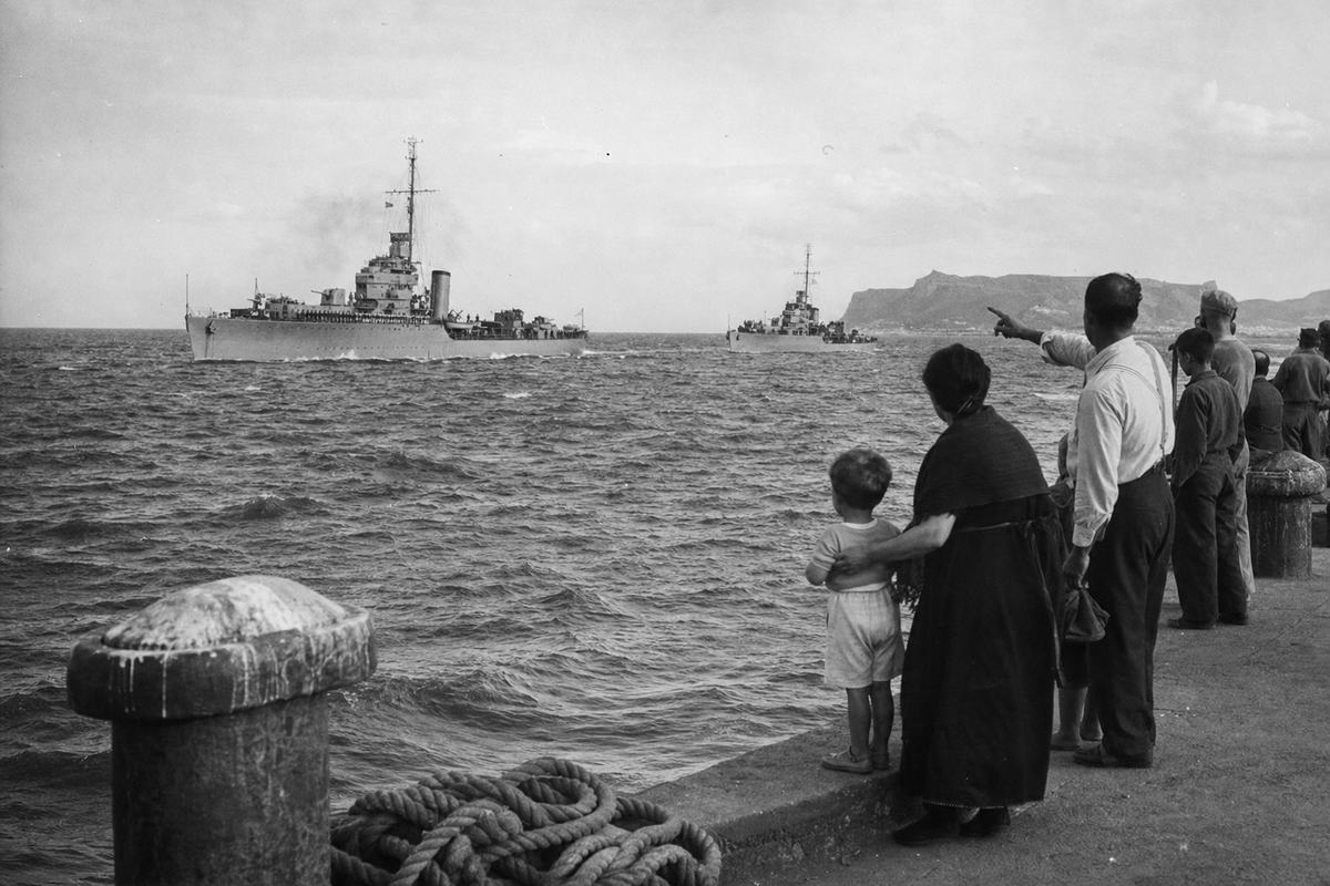 Black-and-white press photograph from late 1938 showing two Royal Navy destroyers lying offshore near a Spanish port (Valencia or Alicante): asymmetrical composition from the quay with mooring bollards in the near left and a coiled hawser; civilians and dockworkers of distinct ages and builds watch the grey hulls from the right edge, one man in suspenders pointing seaward while a woman steadies a child; low swell with whitecaps; distant headland faint in haze; no visible text or ship names. Shot on panchromatic nitrate film, moderate grain and high-contrast sky, Contax II with 85mm lens; slight lens vignetting and salt spray speckling on the negative.