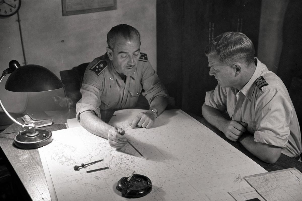 Archival black-and-white interior photograph circa 1940 of a liaison room at the naval base in Cartagena: two officers seated at a scarred wooden table, one Spanish in khaki tunic with rolled sleeves, one British in shirtsleeves; the Spanish officer gestures with a pencil toward an unrolled sea chart showing coastlines and gridlines with labels cropped or too soft to read; dividers, a grease pencil, and an ashtray with a smoldering cigarette sit on the table; wall clock partially cut off; a shaded desk lamp creates falloff across the paper; no legible text visible. Shot on medium-format camera (Rolleiflex 75mm), fine-grain panchromatic film, gentle halation around the lamp, slight edge softness.