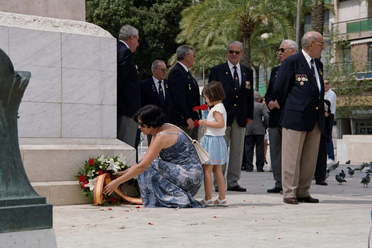 Color photograph from 1998 of Alicante’s postwar memorial during an anniversary commemoration: oblique view with marble steps and low stone plinth partly cropped so any inscriptions are out of frame; veterans in navy blazers with mixed medals stand at ease, a woman in a summer dress kneels to adjust a wreath ribbon while a child holds carnations; scattered petals on paving stones; pigeons at the far edge; bright Mediterranean light with mild shadows; faint salt staining on stone; no text visible. Shot on Fujicolor 200, slight warm cast, 50mm lens, moderate depth of field.