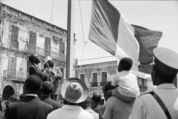 Archival black-and-white press photograph taken on June 20, 1960 in Saint-Louis, Senegal River waterfront square. Shot on 35mm Kodak Tri-X 400 with a Leica M3 and 50mm lens, high noon natural light, hard shadows, pronounced film grain, slight motion blur in the flag and crowd, minor negative scratches and dust specks consistent with archive prints. Asymmetric composition: Léopold Sédar Senghor and Modibo Keïta at left mid-ground, distinct faces and builds, jointly pulling a halyard to raise a new flag; the flagcloth caught mid-lift. Foreground shoulders of onlookers, a straw hat, a child lifted on someone’s shoulders; mid-century suits and dresses; a gendarme partly cropped at frame edge; background shows weathered colonial façades, balconies with peeling paint. No text, labels, numbers, or lettering visible anywhere.