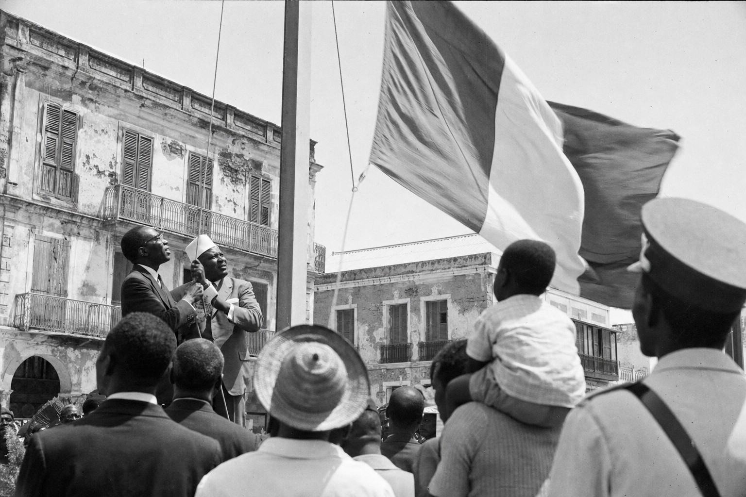 Archival black-and-white press photograph taken on June 20, 1960 in Saint-Louis, Senegal River waterfront square. Shot on 35mm Kodak Tri-X 400 with a Leica M3 and 50mm lens, high noon natural light, hard shadows, pronounced film grain, slight motion blur in the flag and crowd, minor negative scratches and dust specks consistent with archive prints. Asymmetric composition: Léopold Sédar Senghor and Modibo Keïta at left mid-ground, distinct faces and builds, jointly pulling a halyard to raise a new flag; the flagcloth caught mid-lift. Foreground shoulders of onlookers, a straw hat, a child lifted on someone’s shoulders; mid-century suits and dresses; a gendarme partly cropped at frame edge; background shows weathered colonial façades, balconies with peeling paint. No text, labels, numbers, or lettering visible anywhere.