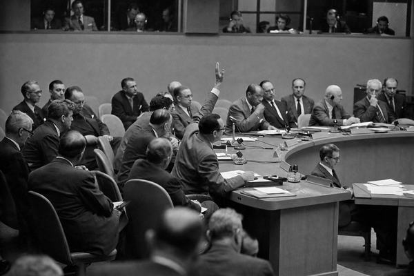 Archival black-and-white photograph, silver gelatin print from a 4x5 negative: UN Security Council session at Lake Success, May 1951, during a vote on a Chapter VII resolution concerning Palestine. Asymmetric composition from the public gallery, focusing on one delegate’s raised hand mid-vote at the horseshoe table; nearby delegates lean toward desk microphones; interpreters visible behind glass; papers and ashtrays scattered on desks. Shot on a Speed Graphic 4x5 with Kodak Super-XX film; moderate grain; slight flashbulb falloff on foreground; shallow depth from a 127mm lens; gentle motion blur on a turning head. No legible text or signage; period furnishings and suits; soft 1950s tonality.