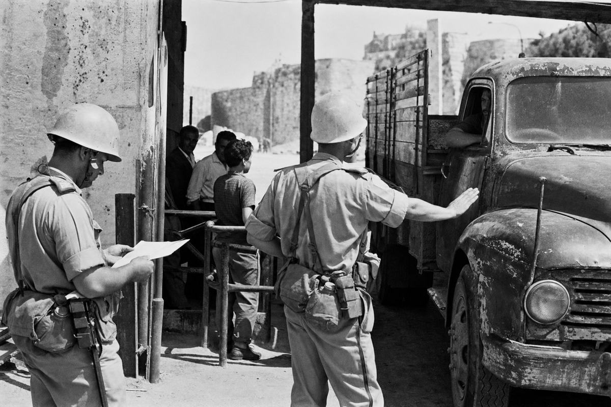 Black-and-white press photograph, mid-1960s: two UN peacekeepers of the Palestine Stabilization Force at the Mandelbaum Gate checkpoint in Jerusalem stop a battered flatbed truck. Off-center frame: one peacekeeper examines a folded paper manifest (text not readable) while the other signals with an outstretched palm; civilians wait by a low turnstile. Light-colored helmets without legible markings; scuffed webbing; dust on boots; patched, dented metal gate; a stray dog in background shade. Shot on a Nikon F with Kodak Tri-X 400 pushed to 800; pronounced grain; high contrast; slight edge vignetting; 50mm lens. No readable signs or lettering in scene.