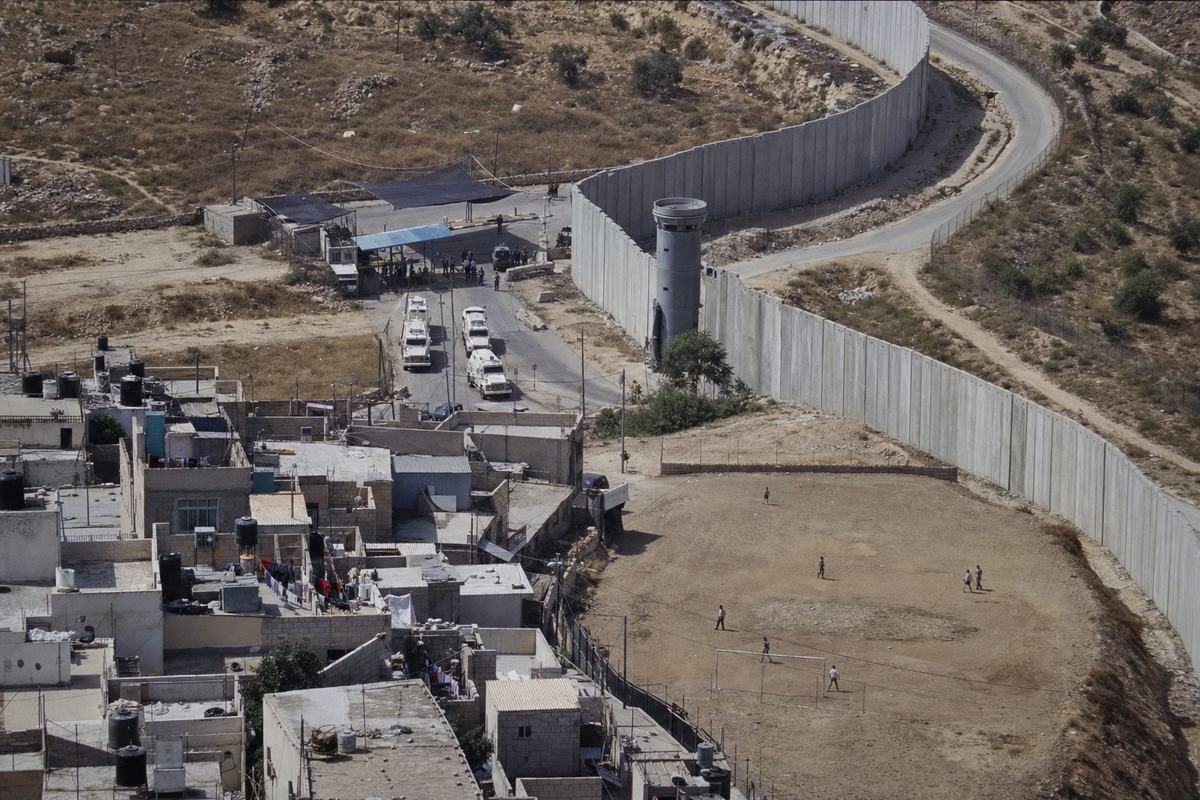 Aerial digital press photograph from a helicopter, early 2003: the separation barrier arcs around the UN trusteeship zone near Jerusalem. Off-center composition: concrete segments curve away from a hilltop watchtower toward an FSP-controlled crossing, where a line of white peacekeeping vehicles (no legible markings) sits in patchy shade; below, rooftops with water tanks, laundry lines, and a dusty soccer field. Captured on a Canon EOS-1D with 70–200mm f/2.8 at 1/1000s, ISO 800; fine high-ISO noise; slight atmospheric haze; mild motion blur at frame edges. No readable text or signage.