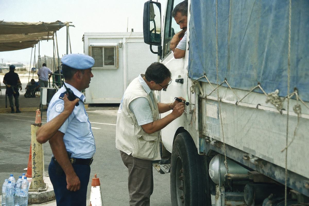 Color photograph on Fujicolor Press 800 film, mid-1990s, at the UN-supervised Ashdod–Jerusalem corridor checkpoint. Asymmetric mid-ground action: a UN administrator in a pale vest inspects a metal cargo seal with a small flashlight while a peacekeeper observes, right hand on a radio; the truck driver, middle-aged and sunburned, leans from the cab window. Scuffed orange cones, frayed rope on blue tarps, a portable inspection trailer and shade tents in background; heat shimmer above asphalt; a cluster of half-empty water bottles by a bollard. Shot on a Canon EOS-1N, 35mm lens; slight greenish cast; visible color negative grain; no legible license plates or signage.
