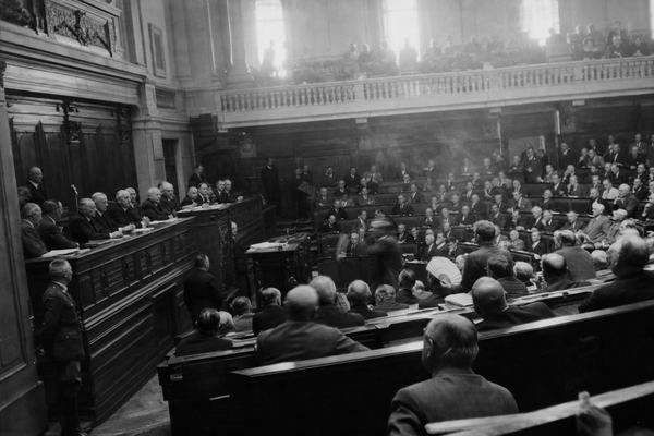 Archival black-and-white press photograph of the joint parliamentary session at Vichy on July 10, 1940; silver gelatin print on Kodak Super-XX film with medium grain and high contrast; candid, off-center view from the right-side gallery looking down with a 50mm lens (Contax II), showing a tense presidium at a wooden dais to the left and crowded benches fanning diagonally across the frame; one deputy mid-gesture blurred, others leaning forward, varied ages and builds, rumpled suits, a few uniforms at the aisle; harsh window light from high clerestory causing slight halation and uneven exposure; handheld camera shake at 1/60 sec; incidental details include handheld fans, cigarette smoke haze, scuffed floorboards, and gallery railings; no visible text or signage anywhere in frame.