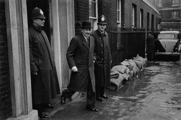 Archival black-and-white press photograph (silver gelatin print), June 1943, outside the doorway of 10 Downing Street, London; Clement Attlee in dark suit and bowler hat steps through the doorway, flanked by two uniformed British police constables; rain-darkened pavement with puddles, wet brickwork, a 1940s saloon car parked upstreet, sandbags piled near a basement window; photographed with a Rolleiflex TLR (75mm lens) at waist level, slight motion blur on Attlee’s stride, moderate Ilford HP3 grain, soft edge falloff and uneven exposure under overcast sky; candid, off-centre framing that crops the doorframe and excludes any visible numerals or signage; natural skin texture, damp coats, no legible text anywhere in frame.