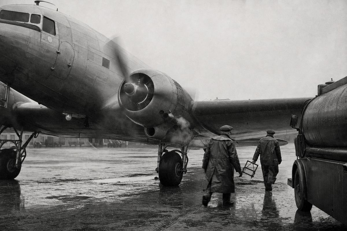 Archival black-and-white photograph (silver gelatin press print), 1943 British airfield (Bristol Whitchurch); a BOAC Douglas DC-3 idles on rain-slick tarmac, port engine turning with propeller blur and exhaust haze; two ground crew in oilskins and flat caps walk beneath the wing, one carrying chocks; a Bedford fuel bowser half-seen at frame edge; shot on a Speed Graphic 4x5 with 127mm lens, shallow depth from flat overcast light, pronounced Kodak Super-XX grain, faint drizzle flare; asymmetrical composition with the nose cropped, aircraft registration and any lettering obscured or out of frame; puddles and tyre tracks in foreground, no legible text visible.