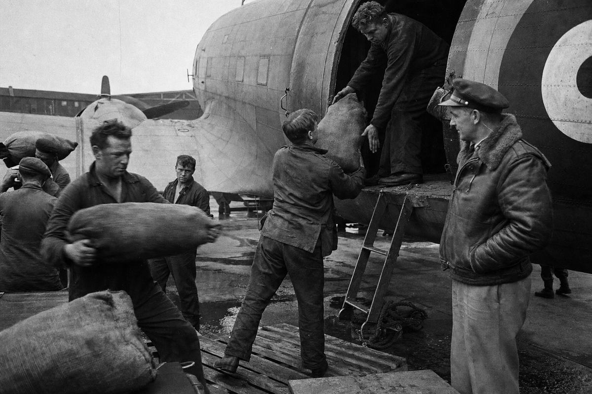 Archival black-and-white press photograph (silver gelatin), 1948 airfield apron at RAF Gatow or Tempelhof during the Berlin Airlift; an RAF Dakota (C-47) stands with cargo door open as ground crew pass hessian coal sacks hand-to-hand; a pilot in a leather jacket watches by the ladder, faces and hands dusted with coal; shot with a Speed Graphic 4x5 using flashbulb fill, Kodak Super-XX film, pronounced grain and high contrast, slight blur on moving sacks; asymmetrical framing with the aircraft tail cropped, hangar structures blurred in background with no legible numbers or signs; damp concrete, scattered ropes and pallets, no text visible.