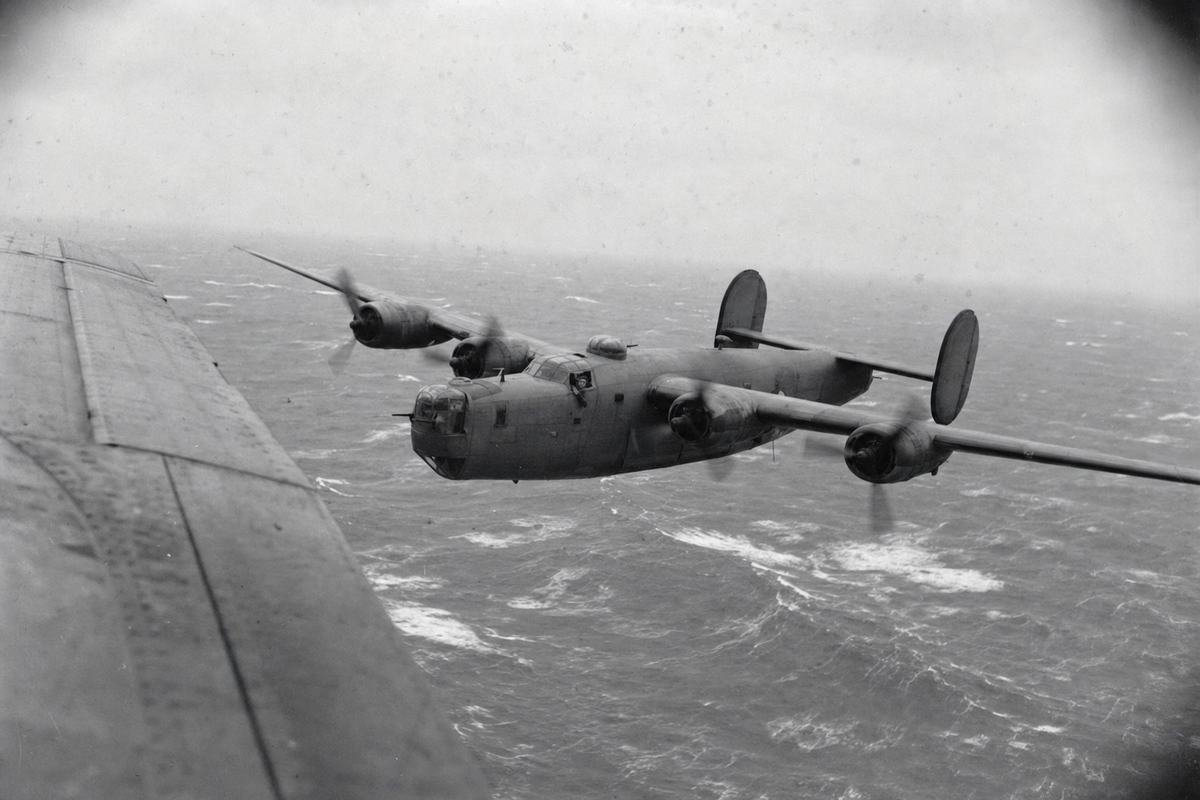 Archival black-and-white photograph taken from another aircraft over the Bay of Biscay, 1943; an RAF Coastal Command Consolidated B-24 Liberator banks low over grey, whitecapped seas, the wingtip and riveted fuselage of the photographer’s aircraft intruding at left; crew figures visible through an open waist window, distinct faces and headgear; handheld Leica III with 50mm lens on Ilford HP3 film, motion blur across propeller arcs, corner vignetting, salt spray spotting on the negative; off-centre framing, squadron codes and any lettering not legible; overcast horizon, no visible text.