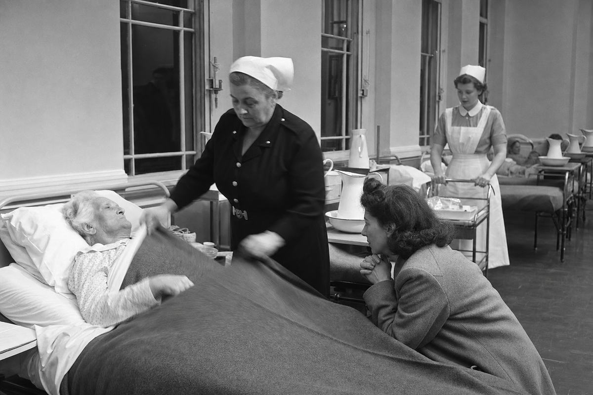 Archival black-and-white press photograph (silver gelatin), interior hospital ward, 1947; a newly opened National Health Service ward with iron-framed beds, white enamel basins, and high sash windows; a matron adjusts a blanket for an elderly patient while a young staff nurse wheels a trolley; a family visitor sits at bedside with clasped hands; shot on a Rolleiflex TLR 75mm with flashbulb bounce, Ilford HP3 film, crisp midtones and mild halation around highlights, slight motion blur in the matron’s hands; candid, off-centre composition; uniforms slightly creased, scuffed linoleum floor; no posters, placards, or legible signage in frame.