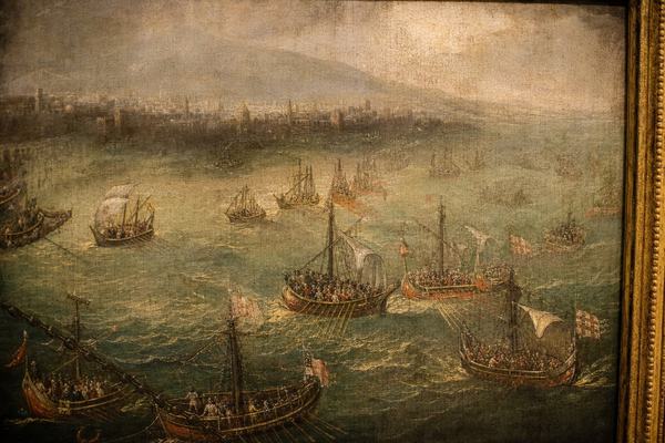Archival museum reproduction photograph of a late-15th-century oil painting on wood panel depicting the relief fleet entering the Golden Horn on April 20, 1453 under passing rain squalls; Venetian and Genoese oared galleys and three Papal galleys surging past the slack harbor chain within the painted scene, chain running diagonally across the lower left; Galata workshop style with compact ship forms, lead–tin yellow wave highlights, verdigris greens in choppy water; visible patina with craquelure and slightly yellowed varnish bloom; narrow gilt frame edge intruding on the right; shot slightly off-center to the left to avoid glare, warm museum lighting, no wall labels or any lettering visible; captured in 2003 with a full-frame digital press camera and a 50mm lens at f/4, shallow depth of field separating the panel from the neutral wall, fine high-ISO noise in darker passages.