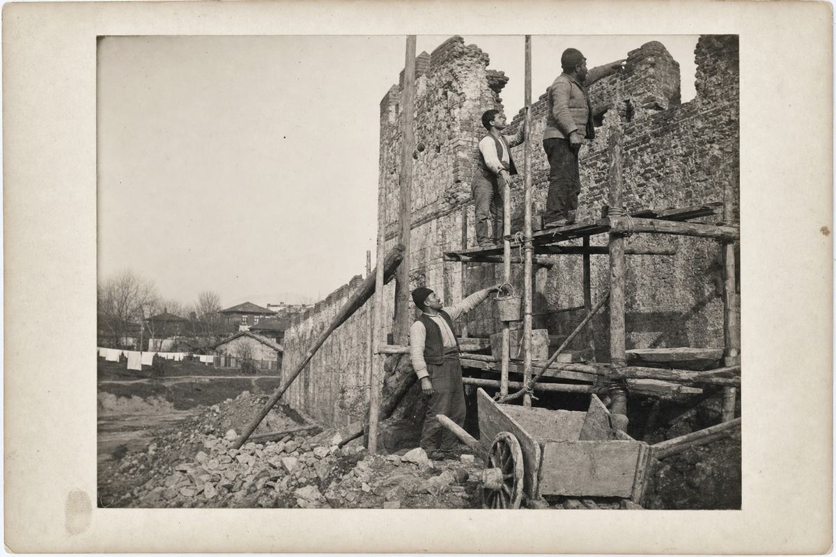 Early 20th-century silver gelatin print on matte fiber paper showing the Theodosian Walls under restoration: timber scaffolding lashed up a crenellated stretch beside a breached tower; three distinct masons at staggered heights—one older with a trimmed mustache handing up a bucket, a lean youth bracing a pole, a stockier foreman pointing toward a joint; foreground rubble piles and a handcart with a cracked wheel; soft winter light with long tonal range and gentle contrast; fine grain with slight edge vignetting and a thumbprint in the lower margin; photographed c. 1908 with a 9x12 cm plate camera on tripod, normal lens; background reveals scattered low houses, a line of laundry, and bare trees beyond the wall; candid, asymmetrical framing with more sky at left; no signage or any lettering visible anywhere.