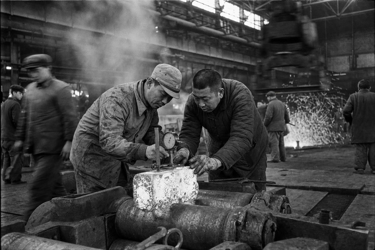 Archival silver-gelatin print, mid-1930s, Shenyang rolling mill; photographed with a Leica II and 50mm lens on Agfa Isopan film; pronounced grain, high contrast, slight motion blur from an overhead crane; asymmetric low angle beside the rolling line; dominant subject a paired team of foremen—one Japanese in grease-streaked coveralls and cap, one Chinese in a quilted jacket—bending over a dial gauge atop a glowing billet; sparks in the rear, steam haze, riveted beams, oil stains on concrete; workers of distinct ages and builds move at different depths; subtle edge vignetting; no legible signage or machine plates visible.