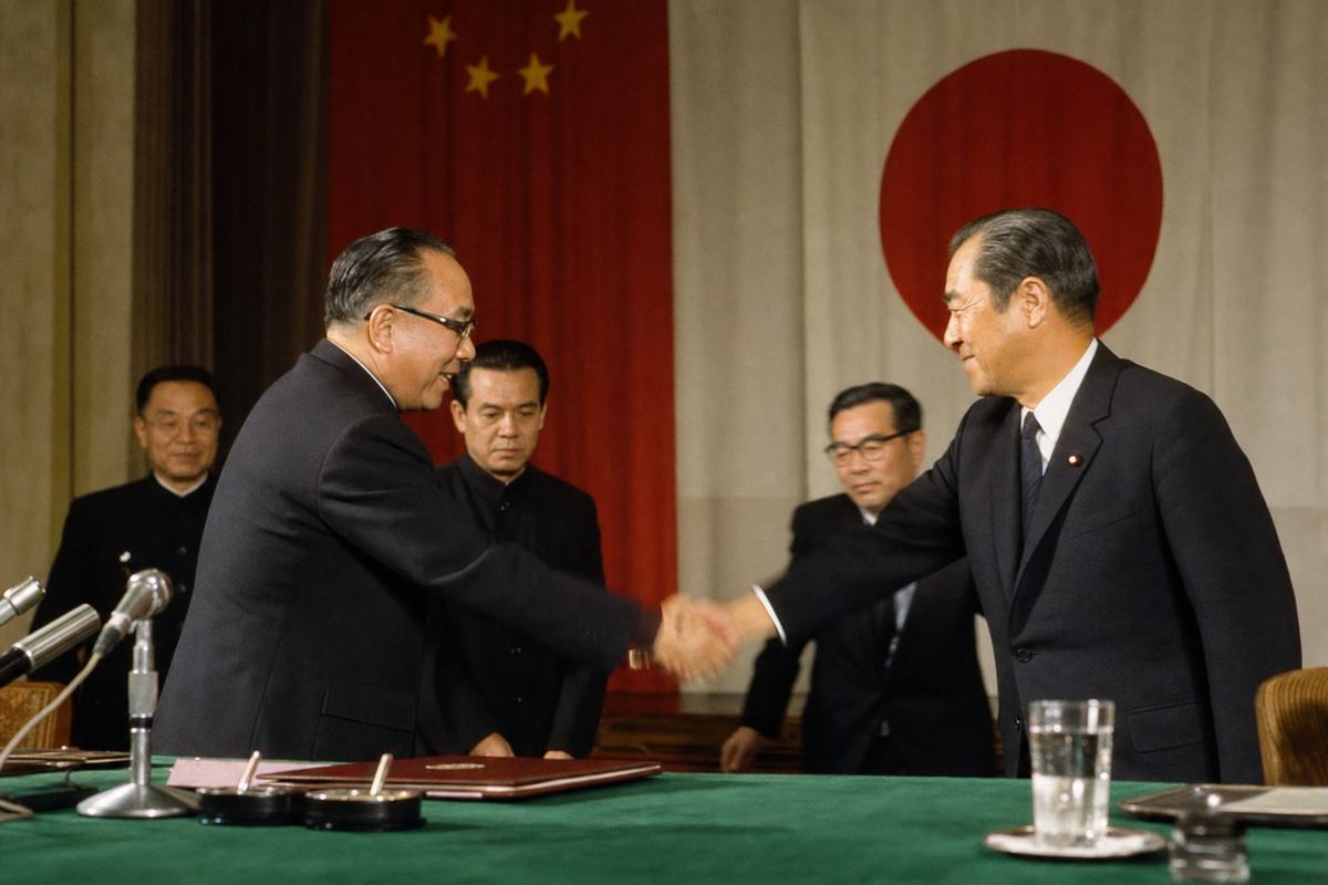 Color photograph on Kodachrome 64, Nanjing, 1965 Normalization ceremony; Nikon F with 50mm f/2 at 1/60, slight motion blur in hands; asymmetric composition from just off the dais; dominant subject the off-center handshake between Premier Zhang Qun and Prime Minister Satō Eisaku across a green baize table; aides of distinct ages and builds at different depths; national flags hang soft in the background; foreground microphone stand and a water glass near the frame edge; warm color cast with deep reds and greens, realistic skin textures; no podium signage or nameplates visible.