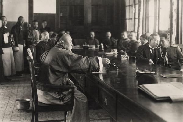Archival indoor silver-gelatin photograph, 1915, Chinese Foreign Ministry in Beijing; taken with a large-format wooden field camera (8x10 glass plate, orthochromatic emulsion), long exposure with slight hand blur and gentle halation around window panes; asymmetric composition from the left rear quarter of a long polished table; dominant subject a Chinese minister in a stiff-collared tunic leaning to reach an inkstand at center while a Japanese envoy in a dark frock coat opposite turns to whisper to a military aide; attendants line the wall in soft focus; worn parquet floor, spittoon near a chair leg, ashtray with a stubbed cigarette, dust motes in slanting light; varied ages and faces with natural skin texture; shallow depth of field falling off toward the far end of the table; no legible documents or signage anywhere.