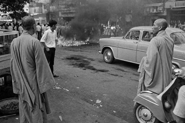 A 1963 press photograph on Kodak Tri-X 400, shot with a Nikon F and 35mm lens, developed as a silver-gelatin print with visible grain, slight highlight blowout, and faint edge vignetting. Captured moments after Thich Quang Duc’s self-immolation at the Phan Dinh Phung–Pasteur intersection in Saigon: asymmetric, candid framing with two robed monks forming a loose semicircle while a young bystander in a white shirt stands at the curb; a scooter and a Peugeot sedan sit halted at angles, heat haze ripples over asphalt darkened by fuel patches. Background shopfronts are soft and partially obscured by smoke; incidental details include a vendor’s pushcart wheel and scattered leaves, no legible signage or text. Natural midday light with shallow contrast typical of Tri-X; slight motion blur in a scooter wheel.
