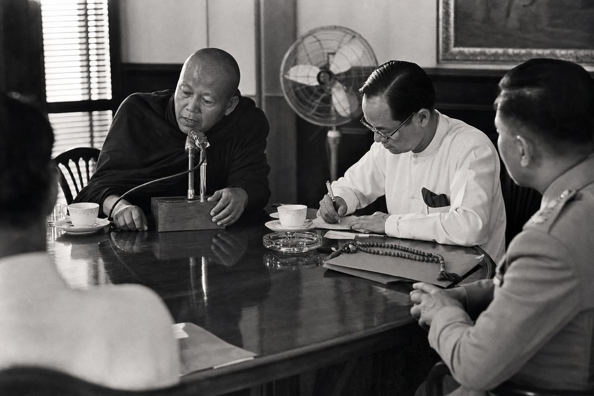 Archival black-and-white press photograph inside the Independence Palace cabinet room, late June 1963; Kodak Tri-X 400 on a Leica M3 with 50mm lens, bulb flash creating hard shadows and falloff; silver-gelatin print with moderate grain and slight corner vignetting. Asymmetric, candid composition: a robed monk co-chair leans toward a wooden microphone base while a bespectacled Catholic lay leader scribbles notes; at the table corner a uniformed officer in profile observes. Teacups, an ashtray, and a loop of prayer beads rest near a manila folder; venetian blinds throw slatted light across the tabletop. Background shows a floor fan and the edge of a framed painting; no legible documents or signage in frame.