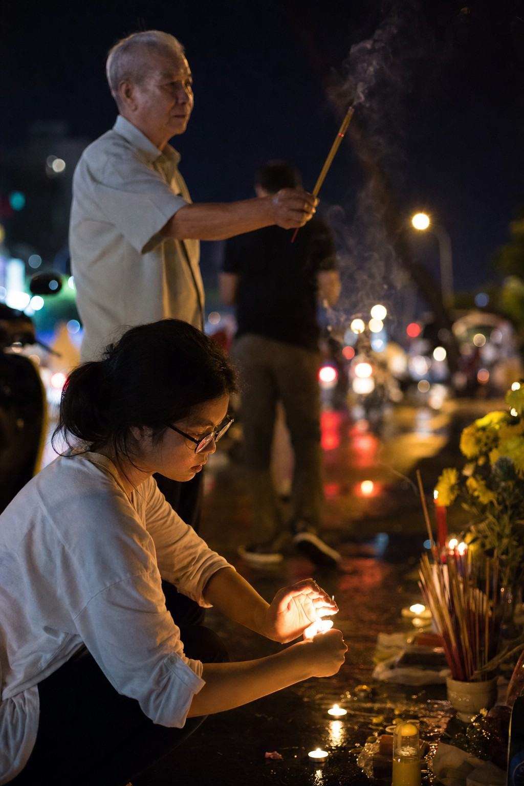Contemporary digital press photograph of a candlelight vigil at the memorial shrine on the Phan Dinh Phung–Pasteur corner in Saigon, 2018; full-frame DSLR (Canon 5D Mark IV) with 50mm lens at f/1.8, handheld at ISO 6400 creating fine high-ISO noise and shallow depth of field. Asymmetric composition: a university student in three-quarter profile shields a small candle flame while an elderly man behind her raises a joss stick; incense smoke lifts in a tall column and drifts into bokeh from passing scooter taillights. Wet pavement reflects warm candlelight; mixed street lighting casts gentle color shifts; background traffic blurred; no legible signs, banners, or text in frame.