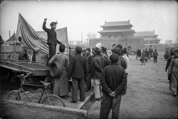 Archival gelatin silver print photograph made in early May 1919 at Tiananmen Square, Beijing; candid, asymmetric composition from a slightly elevated vantage near a wooden handcart; large-format 5x7 glass plate negative with a Zeiss Tessar 135mm lens; soft spring haze, moderate grain, slight edge vignetting, and minor emulsion scratches; dominant subject: a young student in a dark tunic standing on a handcart, arm raised as he addresses telegraphers and railway workers clustered below; distinct faces of mixed ages and builds, natural postures; fabric banners hang slack or turned so no characters are readable; workers with coils of wire over shoulders; foreground bicycle propped against a curb with scattered straw; background Tiananmen gate architecture receding in haze with rickshaws and pedestrians; uneven exposure across the sky; no modern elements, no legible text anywhere.