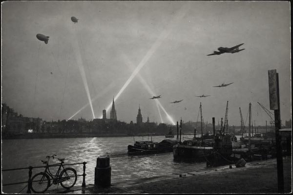 Archival silver-gelatin photograph taken at dusk on 14 May 1940 from the south bank of the Maas in Rotterdam; barrage balloons tethered on long cables overhead and intersecting searchlight beams crossing low cloud; a formation of twin‑engine bombers (period‑accurate Heinkel He 111 silhouettes) banking away over the river; camera: Leica IIIb with 50mm Summar on a small tripod, exposure around 1/10s; Kodak Super‑XX film pushed in D‑76, pronounced grain, slight motion blur on aircraft and searchlight beams, gentle falloff at frame corners; foreground shows a bollard and a dockworker’s bicycle leaning against a railing, moored barges rocking on wind‑ruffled water, gulls in the near distance; skyline off‑center with church towers and cranes in partial silhouette; humid haze softening far buildings; candid, asymmetric framing; no legible text or signage visible; authentic 1940 press photography aesthetic with lightly frayed print edges and faint silvering in the deepest blacks.