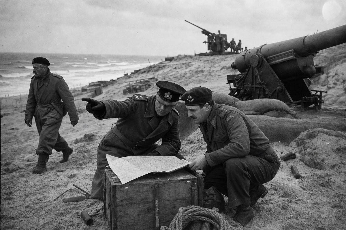 Archival silver-gelatin press photograph made in summer 1940 among the Zeeland dunes: a Dutch officer in a greatcoat and a British major crouched beside an ammunition crate, heads close over a creased map, one gloved hand pointing toward the surf; behind them, the breech of a coastal gun and sandbag revetment, with a Bofors 40 mm silhouette further upslope; wind kicks sand across boots and hems; background sea with whitecaps under low cloud; camera: Rolleiflex Automat 6x6 with Zeiss Tessar 75mm, f/8 for depth; Agfa Isopan F film, medium contrast, fine grain, slight vignetting, a hint of lens flare at upper right; candid, off‑center composition with a sapper in the back left walking past, older and moustached, glancing sideways; bootprints, coiled rope, and half‑buried spent casings visible; no legible insignia or signage.