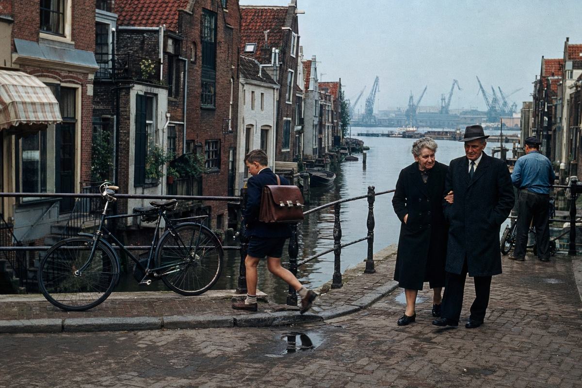 Street‑level color photograph of Delftshaven, Rotterdam, circa 1965: Kodachrome X (ASA 64) transparency with saturated reds and blues and a slight warm cast; Nikon F with 35mm lens at f/5.6; asymmetric composition looking along a narrow canal, brick gabled houses intact and closely spaced; wet cobblestones with small puddles catching sky reflections; two bicycles leaning against an iron railing in the foreground; a schoolboy with a satchel mid‑step on a small bridge, and an elderly couple walking arm‑in‑arm under a dark coat, faces distinct and unposed; distant cranes of Europoort faint on the horizon across the water under a light haze; incidental details of moored skiffs, windowboxes, and a striped awning without any legible text; fine film grain and slight edge darkening typical of period slides.