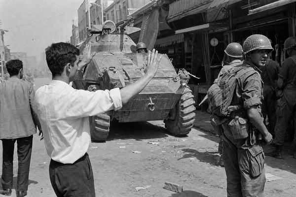 Archival black-and-white press photograph taken in early June 1963 in Tehran near the bazaar district, shot on Kodak Tri-X 400 pushed to 800 with a 35mm lens on a Leica M3; silver gelatin print with visible film grain, slight motion blur at the frame edges, and minor dust scratches from darkroom handling. Asymmetric street-level composition: dominant subject is a young man with rolled-up sleeves standing at left foreground with both palms raised toward an idling armored vehicle; paratroopers in steel helmets and webbing stand along the right side, one glancing back. Background shows worn shop awnings, hanging scales, cracked pavement, and scattered leaflets, none with legible text. Natural midday light with harsh shadows and a low haze of dust; no modern elements or readable signage.