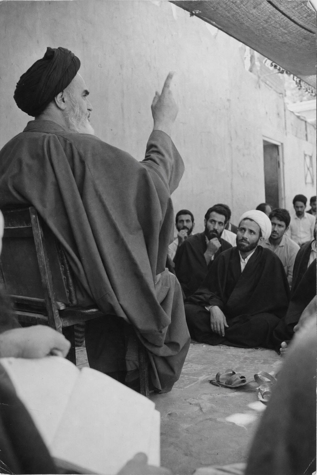 Archival black-and-white press photograph from 1964 in Qom, Iran, captured on Kodak Tri-X 400 with a 50mm lens on a Nikon F; silver gelatin print on matte fiber paper with soft mids, moderate grain, and a faint corner dog-ear from album storage. Asymmetric courtyard composition from slightly behind and below the speaker: dominant subject is Ayatollah Ruhollah Khomeini in three-quarter profile lifting his right hand as he addresses a small gathering. Foreground shows a student’s notebook edge and worn sandals; nearby clerics listen with distinct faces and postures. Bare plaster wall and a sagging fabric canopy create mottled shade; no banners or readable text anywhere.