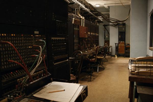 1988 color photograph in a museum study room of a preserved 1930s Bureau wire room re-creation: rows of vintage switchboards, operator stools askew, cable looms sagging along ceiling racks, paper trays with curled edges on a side table. Shot on Kodachrome 64 with a Nikon F3 and 35mm lens at f/4; natural museum can-lights create soft shadows and slight tungsten warmth. Fine, realistic film grain; slight falloff at frame edges. Asymmetric composition with the nearest switchboard half-cut in the left foreground and a narrow aisle receding diagonally. Dust motes in light, scuffed linoleum, and a forgotten pencil on a clipboard. No text or labels visible anywhere.