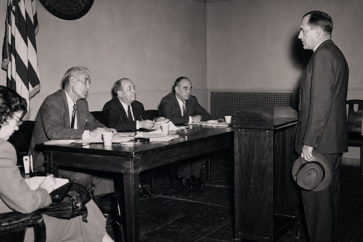 Circa 1952 black-and-white silver-gelatin press photograph of a federal loyalty board hearing: three panelists at a long wooden table under harsh flashbulb light, a mid-40s federal employee stands at a side lectern gripping his hat brim. Shot on a 4x5 Graflex Speed Graphic with single-bulb flash; crisp central detail with falloff at corners, high contrast and slight halation around highlights. Asymmetric framing with the witness off to the right; coffee cups, scattered pencils, and scuffed linoleum add incidental detail. A U.S. flag’s fringe and a wall seal are partially visible but cropped so no lettering is readable. Faces distinct and unposed; a stenographer leans in from the left margin. No legible text anywhere.