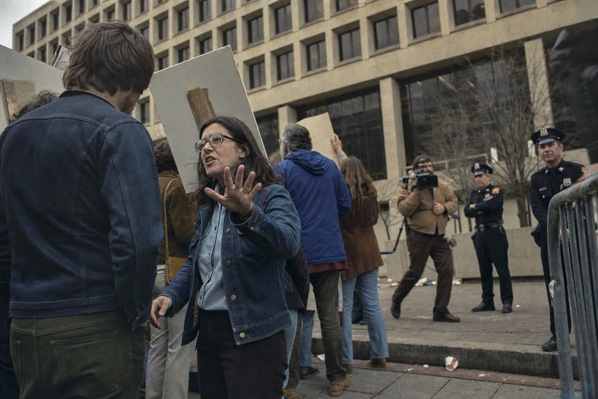 Mid-1970s color photograph of demonstrators outside a federal headquarters building with modernist concrete grid facade: a woman in a denim jacket raises an open palm while speaking to a younger man; several placards are visible from the back or edge-on only. Shot on Ektachrome 100 with a Nikon F2 and 50mm lens; slightly warm overcast light, moderate grain. Police officers stand near a barricade at the frame edge; a press photographer with a shoulder-mounted camera is blurred mid-step in the background; litter and a crushed paper cup near the curb. Asymmetric, low-angle composition that avoids any signage; no legible text on placards or building.