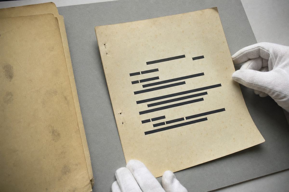 1988 archival object study, color flat-lay of a declassified Bureau memorandum page on gray foam board with white cotton gloves lightly holding a corner; visible paper foxing, staple holes, and solid black redaction bars but no readable characters. Shot on Ektachrome 100 with a 60mm macro lens; soft top-light on a copy stand creates gentle shadows and slight vignetting. A manila folder sits partly in frame with its tab turned away; edges show wear and faint fingerprint smudges. Composition diagonal from lower left to upper right. No text or numbers legible anywhere.
