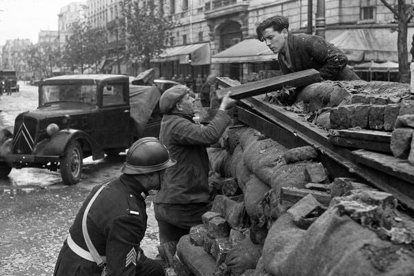 Archival black-and-white silver gelatin press photograph, mid-June 1940, street-level on Boulevard Saint-Michel, Paris. Primary subject: a sandbagged barricade built from turned tram rails, cobblestones, and timber, with a Garde républicaine sergeant crouched left, a middle-aged civilian in a flat cap handing up a rail tie, and a younger student with dust on his jacket peering over the top. Period vehicles halted in the midground (a dark Citroën truck angled across the lane), scattered debris and broken glass along the curb. Overcast sky; damp pavement; leaves stuck to sandbags. Shot on a Leica IIIa with 50mm Elmar at roughly f/4, 1/50s on Agfa Isopan film; moderate grain, slight motion blur on the civilian’s hand, soft vignetting at the corners. Asymmetric composition with the barricade rising from lower right to center; background facades and awnings present but without readable signage; no text or lettering visible; realistic skin texture and period clothing; photojournalistic framing.