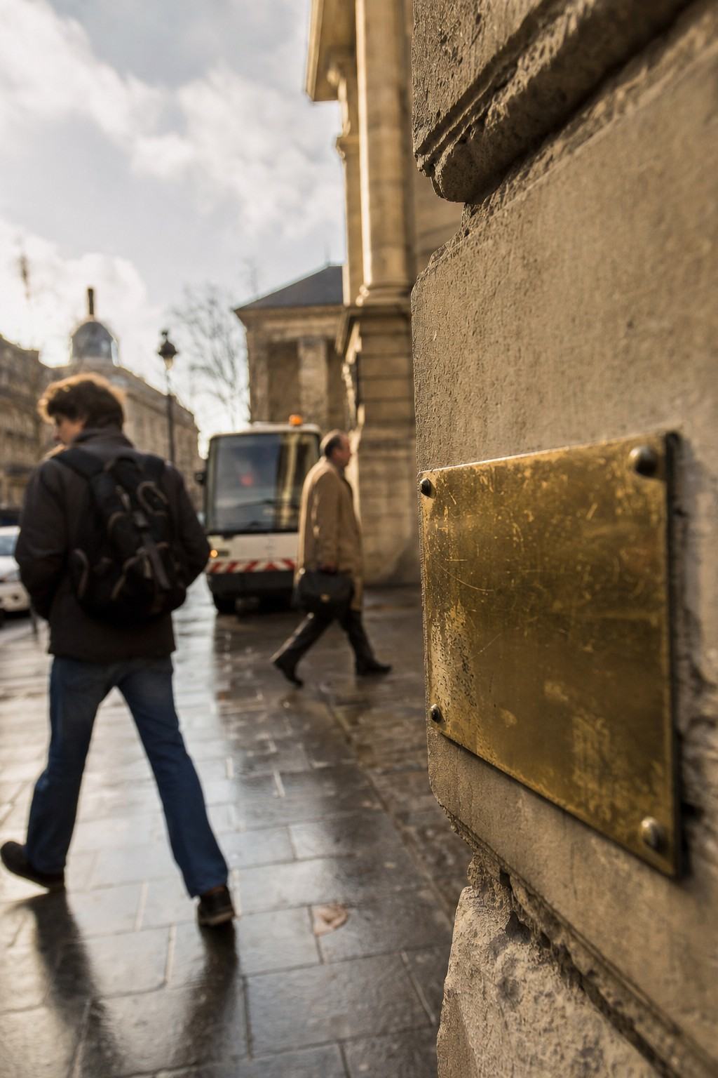 Contemporary color digital press photograph, early 2000s, taken near the Sorbonne in the Latin Quarter. Primary subject: a small brass plaque mounted waist-high on a stone wall, photographed at a shallow oblique angle so its face catches light but lettering is not visible; patina and edge wear evident. Midground: a municipal van idling at the curb; two pedestrians pass in opposite directions, one with a backpack, one older in a tan coat; wet pavement reflecting muted sky. Shot on a Canon EOS-1D with a 35mm f/2 lens at wide aperture; shallow depth of field isolates the plaque edge, background softly blurred; slight high-ISO noise in shadows; warm color cast from late-afternoon light. No readable text anywhere in frame.