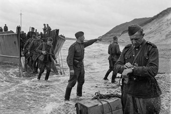 Archival silver-gelatin photograph, August 23, 1945: Soviet infantry disembarking from U.S.-built landing craft with lowered ramps onto the grey pebbled beach at Rumoi, Hokkaido, under a low overcast; surf churning around boots; one officer in a field tunic and side cap points men toward the dunes while another checks a pocket watch; incidental crates and a coiled hawser at the waterline; captured with a 35mm rangefinder (Leica III-type), 50mm lens; moderate grain, slight edge vignetting, salt spray specks on the lower right, uneven exposure near the horizon; candid, off-center framing from a crouched position close to the surf; no visible text or legible hull numbers anywhere in frame.