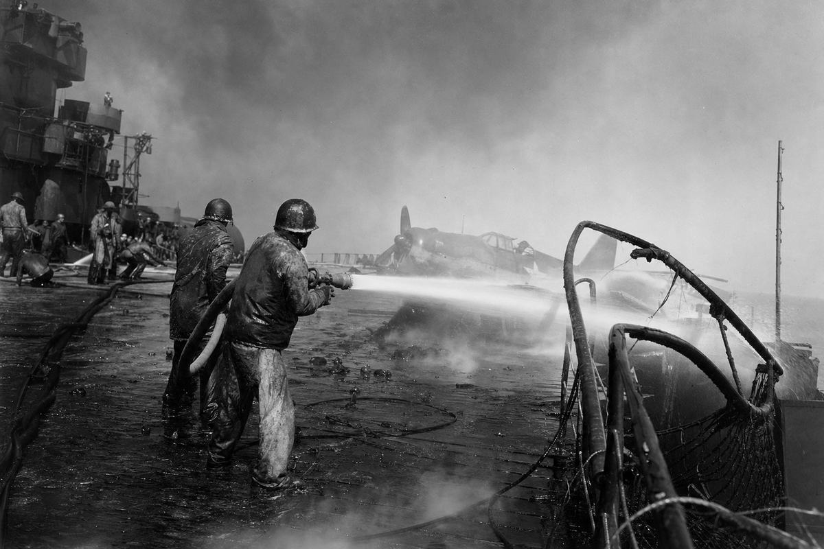 Archival U.S. Navy black-and-white press photograph, June–July 1945: damage-control crews on the flight deck of an Essex-class carrier off Okinawa fighting deck fires; hoses arc foam across scorched plating; a bent catwalk and warped railing frame the right edge; billowing smoke partially obscures a halted aircraft; captured on a 4x5 Speed Graphic with 127mm lens; high contrast, visible soot on negatives, slight motion blur in the hose stream, salt spray halation at the bottom edge; oblique composition from knee height; no legible ship name or hull number visible anywhere.