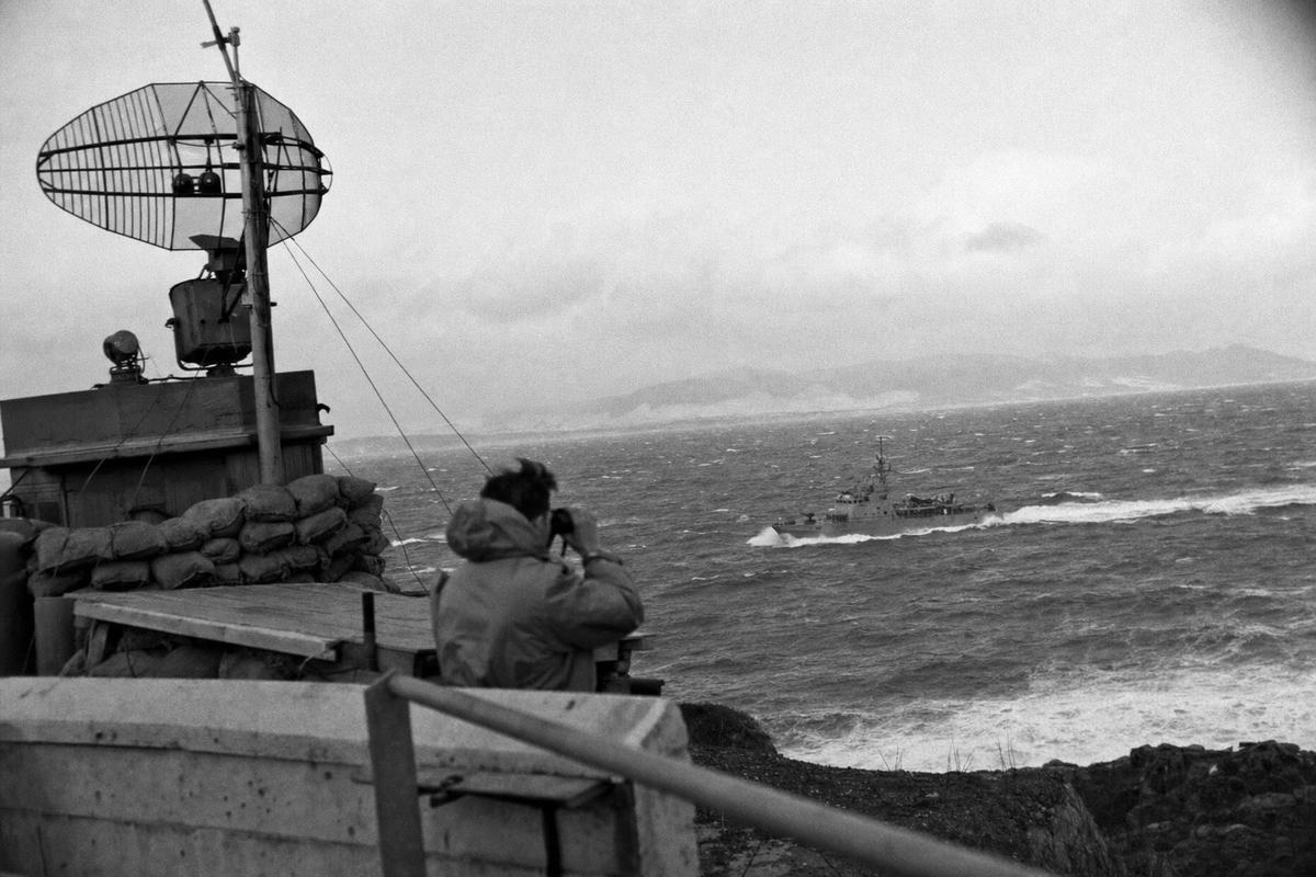 Black-and-white photograph, circa 1964: view from Aomori’s Cape Tappi over the Tsugaru Strait on a gusty day; a sandbagged watchpost and radar mast occupy the left foreground while a patrol boat cuts across whitecaps mid-frame; a coast guard crewman with binoculars stands half-turned, parka hood whipped by the wind; shot on Kodak Tri-X 400 with a Nikon F and 105mm lens; pronounced grain, high contrast, salt mist softening distant shoreline, slight vignetting; off-center horizon and tilted foreground railing; no legible signage or markings.