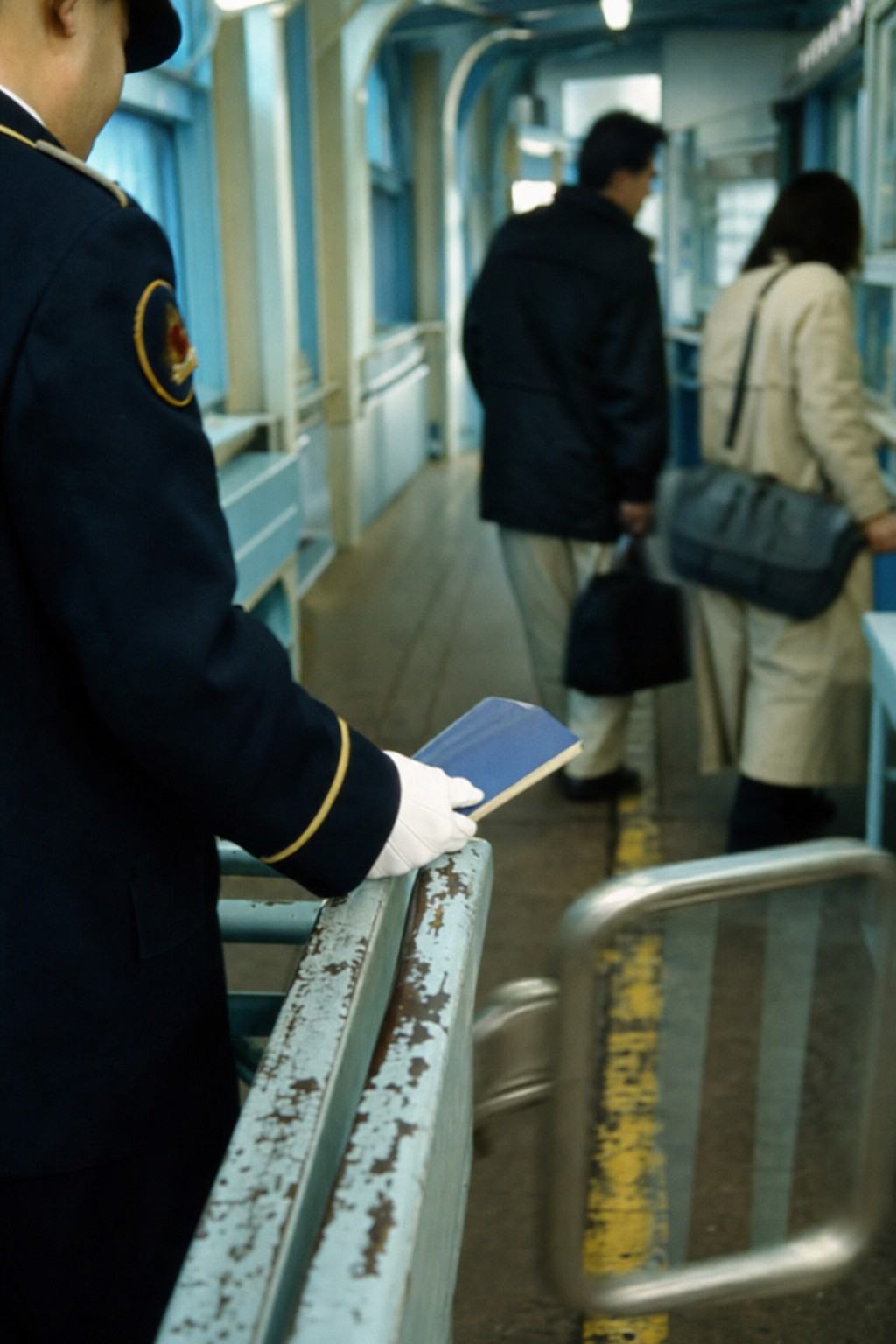35mm color photograph, 1999: Hakodate ferry terminal checkpoint under the Blue Pass corridor; close-up of a uniformed officer’s gloved hand holding a blue travel booklet against a scuffed metal rail, with two travelers waiting out of focus beyond; cool fluorescent cast typical of Fujicolor 400, slightly desaturated blues and greens; shallow depth of field from a 50mm lens, slight motion blur in a swinging gate; worn yellow floor line with chips and shoe scuffs; candid, off-axis framing; no legible text anywhere in frame.
