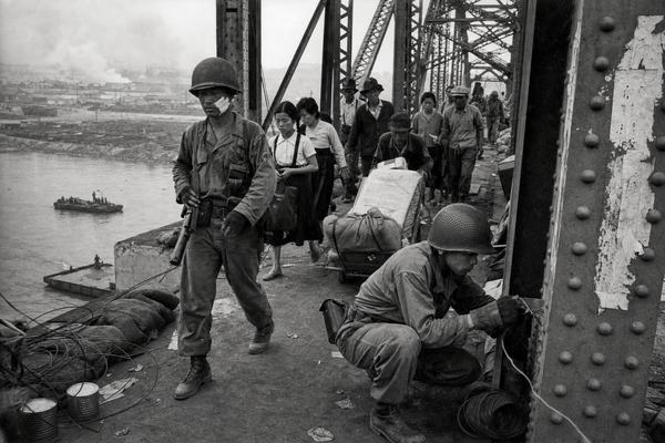 Archival silver-gelatin press photograph taken on the morning of June 28, 1950 on the Hangang Bridge in Seoul; captured with a Graflex Speed Graphic using a 127mm lens on Kodak Super-XX sheet film, processed in D-76. Grain is pronounced with high contrast and slight halation around bright highlights; faint motion blur in marching figures; uneven edge exposure typical of press negatives. Asymmetric, low-angle composition from the south abutment catwalk. Dominant subject: a ROK Army engineer in a steel helmet crouches to tie detonation cord to a riveted girder, gloved hands taut. Behind him, distinct faces of civilians and 5th Division troops stream past: a schoolgirl clutching a satchel, an older man pushing a handcart piled with bedding, a soldier with a bandaged cheek gripping his rifle. Smoke haze softens the far-bank railyard; two river barges drift beneath the truss. Sandbags, coiled wire, scattered ration tins on the deck; torn posters on a pillar with no readable text. No modern vehicles or signage; period uniforms and equipment authentic.