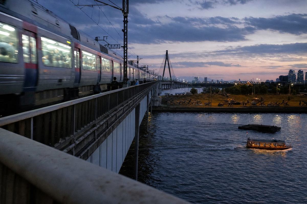 Contemporary digital press photograph taken at dusk in 2015 from the pedestrian deck of the Hangang Bridge; full-frame DSLR with a 24mm wide-angle lens, ISO 3200, handheld, visible high-ISO noise and slight motion blur. Asymmetric framing along the safety railing with leading lines. Dominant subject: a commuter train streaks across the adjacent span, lit windows revealing seated passengers as soft silhouettes; below, a small tour boat and a low barge cut wakes across the river. Background holds the rebuilt bridge pylons and skyline; on the far embankment a small outdoor memorial displays rusted tank obstacles as silent forms. Cool blue-hour color, reflections trembling on the water. No readable text or logos in frame.