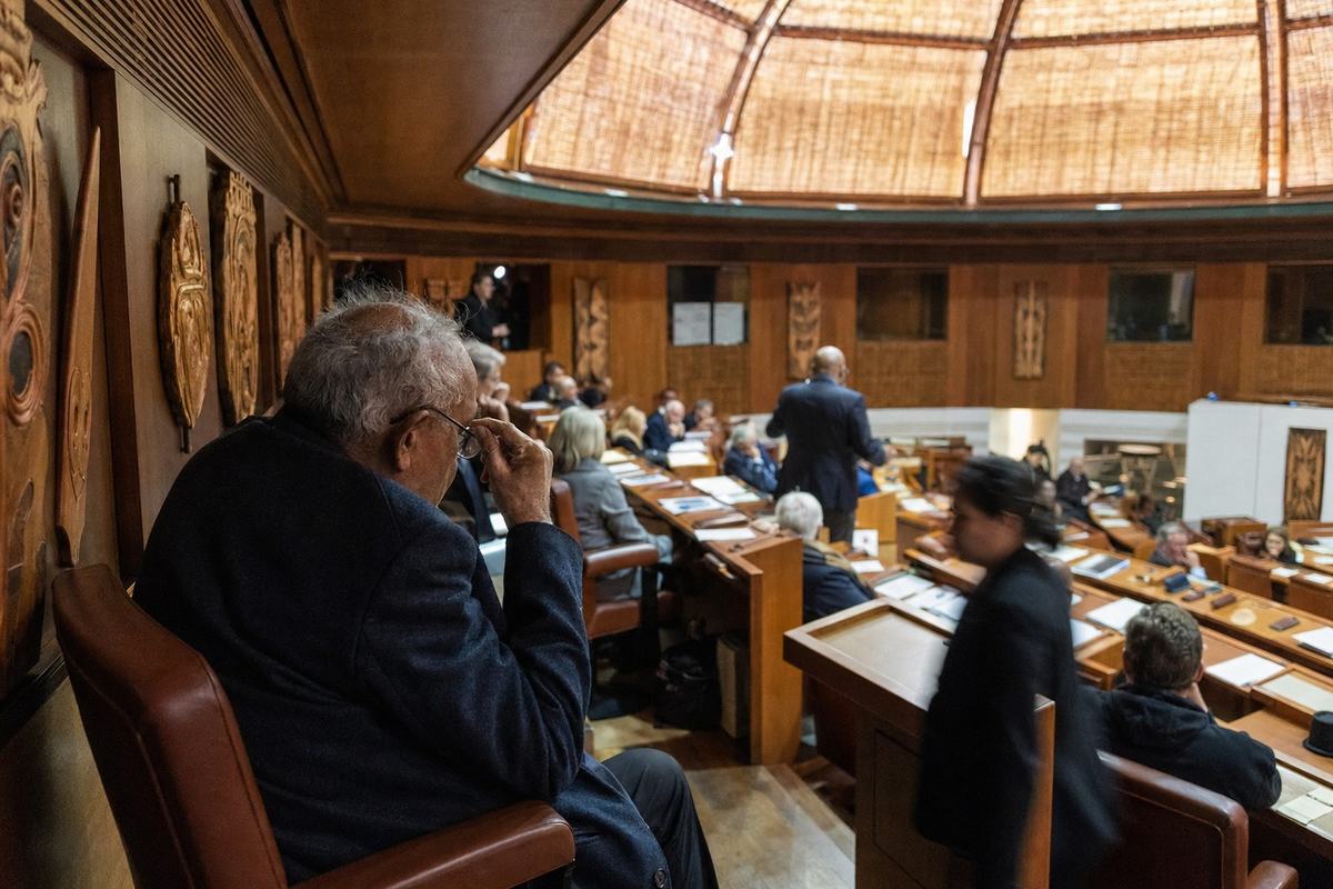 Contemporary digital press photograph inside the House of Accord chamber in Warrane during an anniversary sitting; shot from the side balcony with a 35mm lens on a full-frame DSLR; an Elder seated in the foreground adjusts glasses while a Member rises to speak two rows below, both in natural posture; carved nation shields line the timber walls, woven panels and skylight diffuse warm light; papers and folders lie face-down on desks to avoid legible text; ushers move along the aisle with slight motion blur; high ISO (3200) noise visible in shadow areas, shallow depth of field isolates the central gesture; asymmetric framing with incidental details like scuffed floorboards and worn armrests; no banners or visible lettering