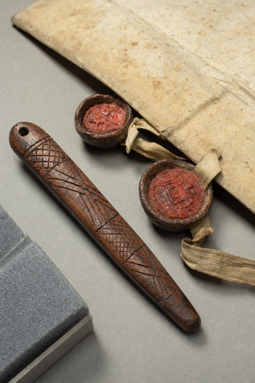 Museum object study photograph on a neutral tabletop: a carved wooden message stick with cross-hatched motifs lies diagonally, beside the lower margin of a parchment compact showing two red wax seals on ribbon; composition cropped to exclude any written text, focusing on seals, ribbon, and carved surfaces; raking light reveals tool marks, patina, and fine dust; slight edge fraying and foxing on parchment; photographed with a 50mm macro lens, shallow depth of field and gentle fall-off; grey archival foam cradle visible at one edge, no display labels or signage
