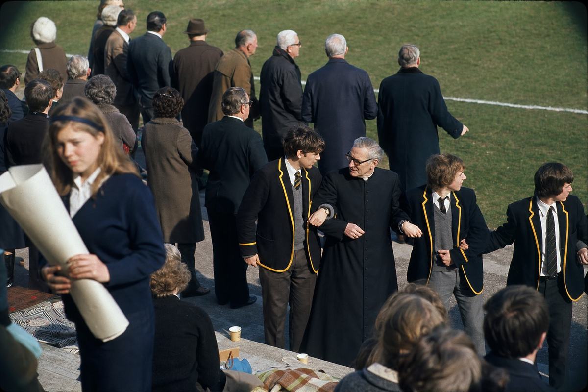Color photograph, June 1976, Kodachrome 64 slide with warm cast and saturated primaries; shot from halfway up the east stand with an 85mm lens at f/2.8 for shallow depth of field. Dominant subject is a line of student marshals linking arms at the touchline, one turning his head to confer with a grey‑haired priest in a dark cassock. Behind them elders in heavy coats cluster near the chalked sideline; foreground shows a girl in a school cardigan gripping a rolled poster with the blank side outward. Concrete terraces dotted with paper cups and blankets; bright winter sun throws hard shadows across the grass; vignetting at frame corners; no legible banners or text.