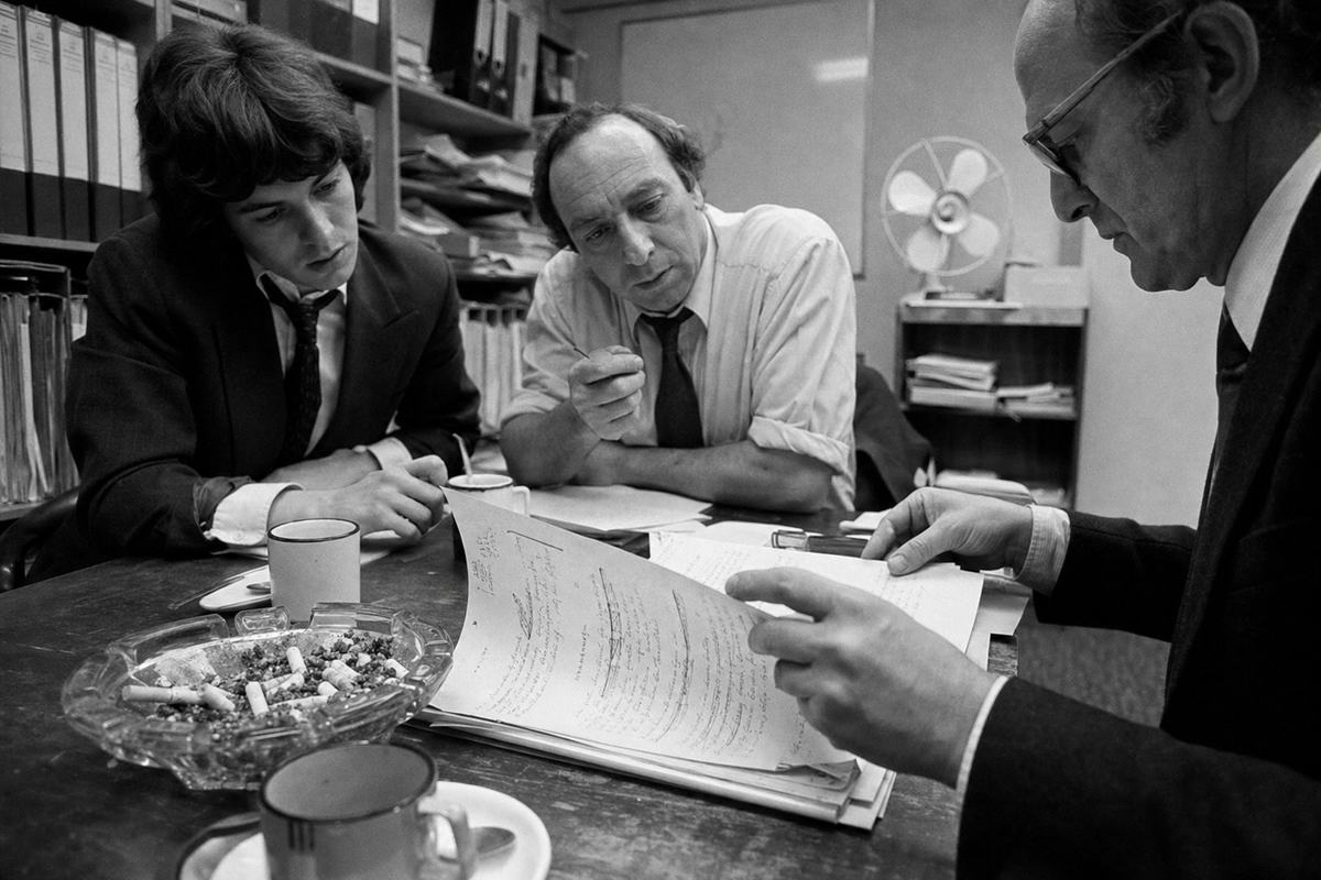Archival black-and-white photograph, June 1976, silver gelatin print from 35mm Ilford HP5 negative; moderate grain, high contrast, and slight lens flare from overhead fluorescents. Cramped government conference room off Market Street; dominant subject is a pair of hands turning a marked-up set of minutes on a scuffed wooden table beside an overfull glass ashtray and enamel teacups. Three negotiators of distinct ages lean in: a student in a blazer with a loosened tie, a middle‑aged civil servant with rolled sleeves, and a church lawyer with wire‑rim glasses. Background shelves with ring binders and a tilted desk fan; no legible document text; captured with a 35mm lens at close distance, slight perspective distortion.