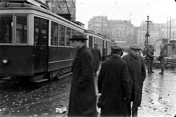 Archival silver-gelatin press photograph taken in Berlin’s Potsdamer Platz during the May 1933 general strike; idle electric trams sitting on wet tracks with trolley poles under slack catenary; a few pedestrians in worn 1930s coats step around puddles; two Reichswehr soldiers stand at a distant checkpoint near sandbags, rifles slung, faces distinct; overcast sky softens contrast; cobblestones dark and littered with leaves and a crushed cigarette pack; captured with a Leica II and 50mm Elmar from street level, slight motion blur on one passerby, medium grain and mild vignetting from aging nitrate negative; asymmetrical, candid composition with a tram car occupying the left third and background buildings kept soft so any signage is unreadable; scanned in the late 1990s from a newsroom print with light edge wear and dust marks.