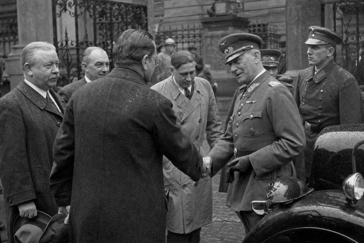 Archival silver-gelatin press photograph outside the Presidential Palace in Berlin on 5 May 1933; candid mid-handshake between ADGB and Christian trade-union leaders and a uniformed Reichswehr major in peaked cap and gloves; the principal hands meet off-center while aides and a junior officer look on from differing heights and builds; cobblestones and a dark state limousine frame the foreground, raindrops speckle coats; a shallow group of onlookers half-hidden behind a wrought-iron gate; captured with a Leica II 50mm at waist level in natural midday light, moderate grain, slight lens flare on metal insignia; background facades rendered soft so any signage is unreadable; asymmetrical, candid framing with the handshake and a turned profile as the dominant gesture; scanned in the 1990s from an original agency print with a light crease at one corner.