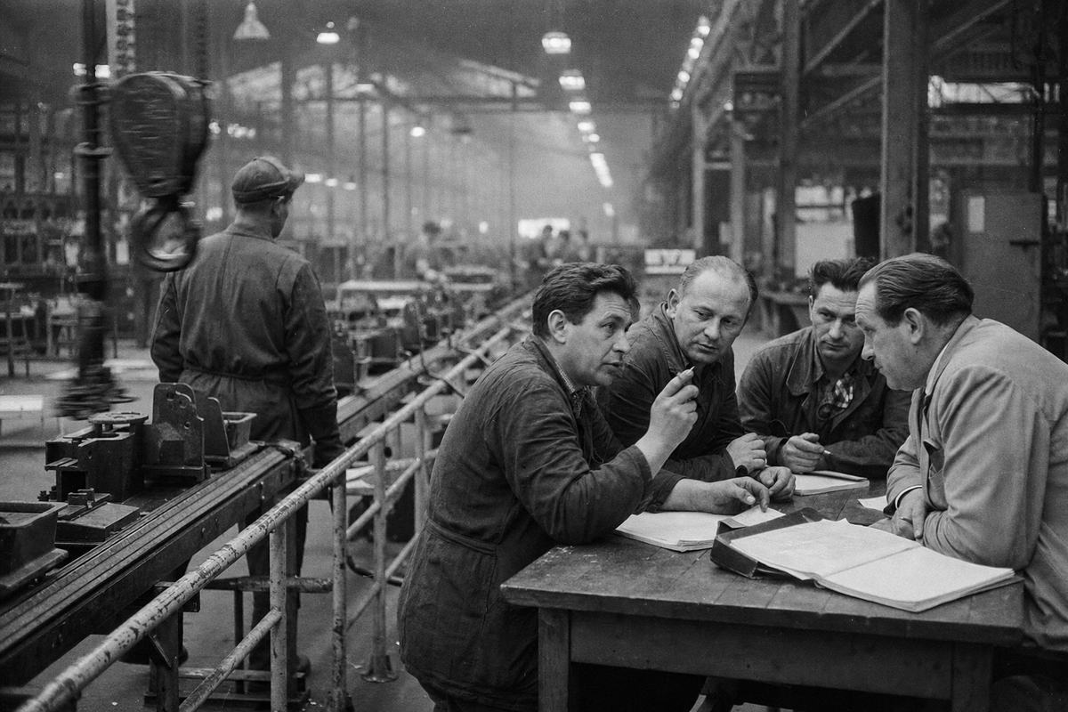 Black-and-white factory-floor photograph of a works-council session in the Ruhr, circa 1957; a trestle table beside an assembly line hosts three elected shop stewards and a plant manager mid-discussion, with papers and a grease-stained ledger open; one steward gestures with a pencil while the manager leans forward, sleeves rolled, faces distinct and lined; conveyor tooling and hanging lamps recede into a hazy background, safety railings scuffed; overhead industrial lighting yields flat highlights; shot on Kodak Tri-X 400 with a Leica M3 and 50mm Summicron, visible grain and slightly blocked shadows, motion blur in a swinging hook; asymmetrical, candid composition with the table occupying the right third; no legible placards or labels; archival scan shows edge rebate and dust specks.