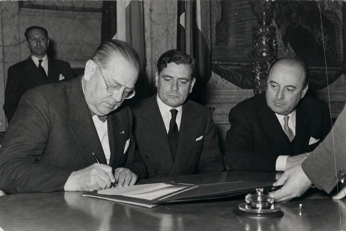 Archival black-and-white press photograph of the 1954 Brussels signing of the European Labor Charter; side-angle view of a Belgian delegate leaning in to set pen to paper while a German and an Italian delegate watch, each face distinct and of different build and posture; a clerk’s hand slides the folder forward from the edge of frame; flags and marble molding blur in the background; flashbulb from a Graflex Speed Graphic creates specular highlights on the tabletop and a mild shadow halo; 127mm lens, high acutance and fine-to-medium grain; papers angled so that any text is out of focus and unreadable; asymmetrical, candid composition with the writing hand as the dominant action; scanned from a newsroom print with slight silvering at dark edges.
